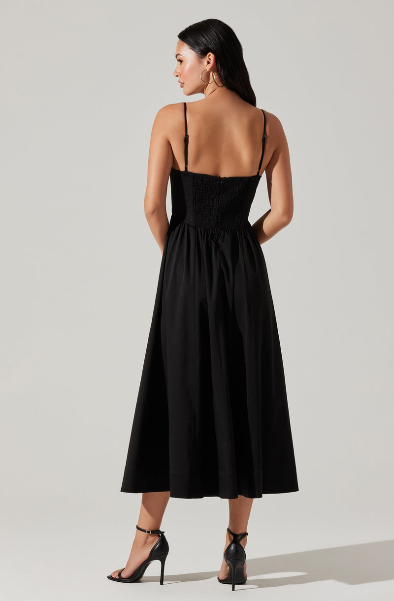 A woman is wearing a black spaghetti strap dress with a fitted, ruched bodice and an elastic waistband. The dress extends to a midi length with a flowy, gathered skirt. She pairs the dress with black high-heeled sandals that have ankle straps.