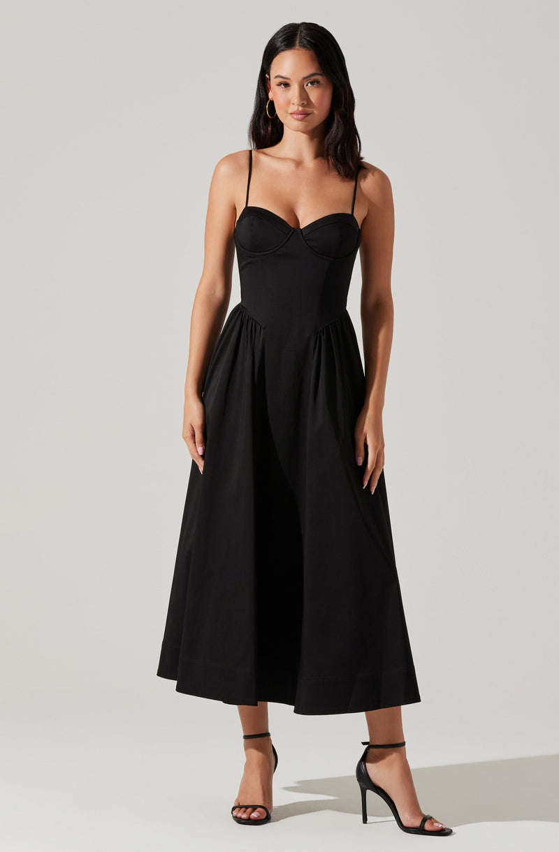 A woman is wearing a black dress with a fitted, strapless bodice and thin spaghetti straps. The dress has a high waistline and a full, ankle-length skirt with gathered detailing. She is also wearing black high-heeled sandals with ankle straps.