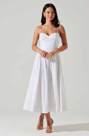 A woman is wearing a white dress with thin spaghetti straps, a fitted bodice, and a full-length skirt that reaches her ankles. She is also wearing nude-colored high-heeled sandals.