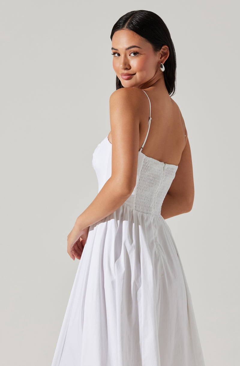 A woman is wearing a white dress with thin spaghetti straps, featuring a smocked bodice and a flowing, gathered skirt. The dress appears lightweight and is styled for warm weather or a casual occasion.