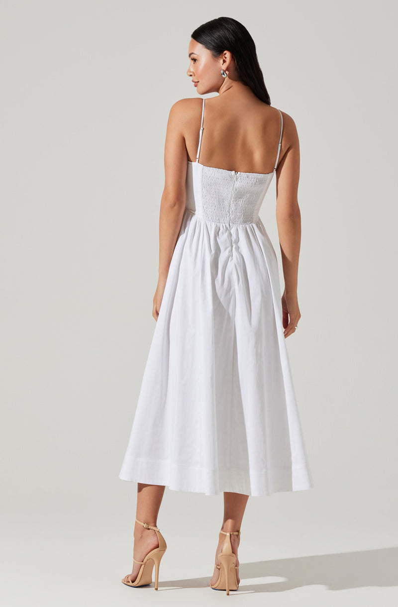 A woman is wearing a white, sleeveless dress with thin spaghetti straps. The dress features a ruched, smocked bodice and a high-waisted, full skirt that falls to mid-calf length. She is also wearing beige high-heeled sandals with ankle straps.