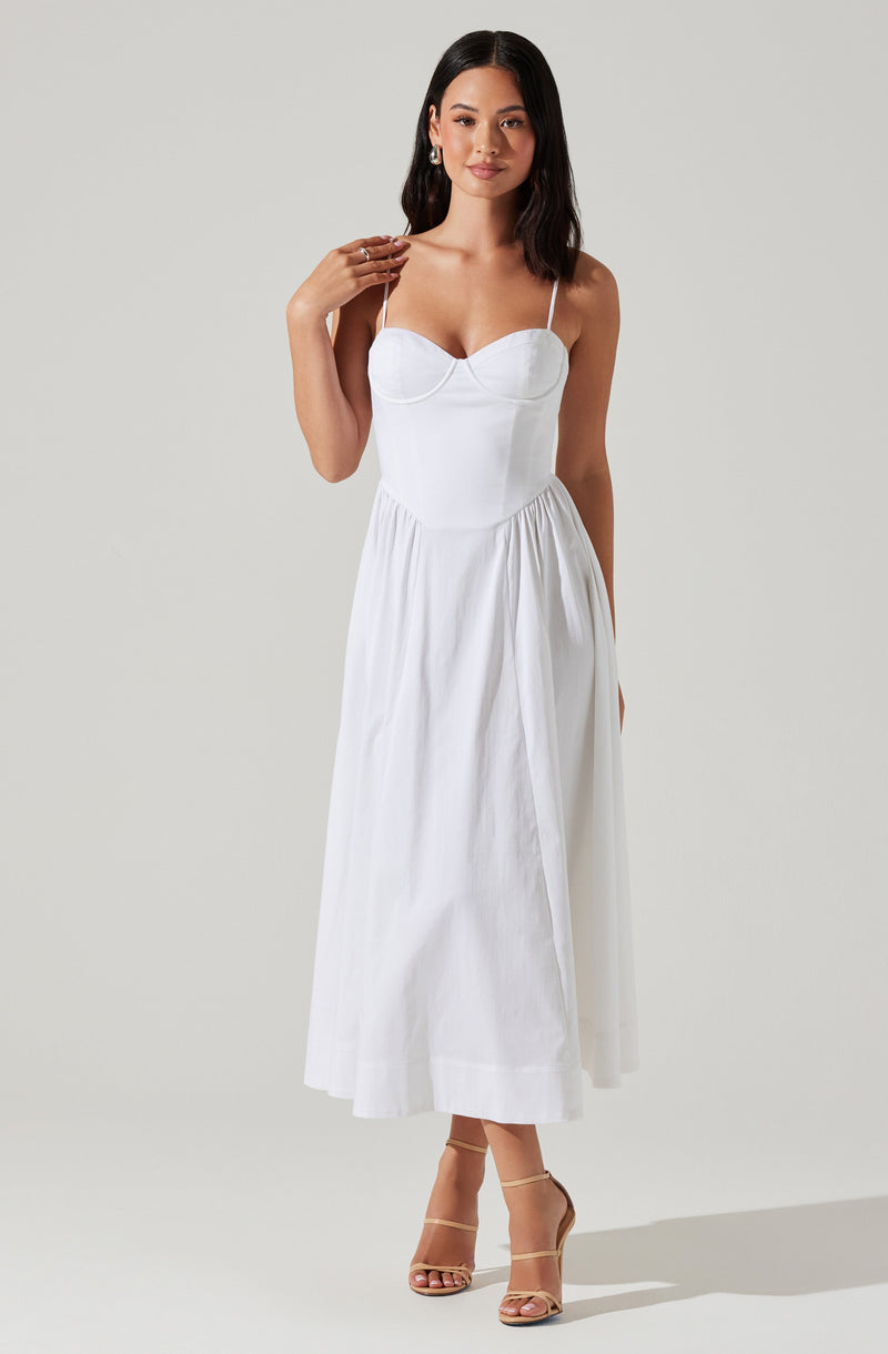 A woman is wearing a white, sleeveless, maxi dress with spaghetti straps and a fitted bodice. The dress has a front seam detail at the bust and a gathered skirt that falls to her ankles. She is also wearing strappy, high-heeled sandals in a neutral color.