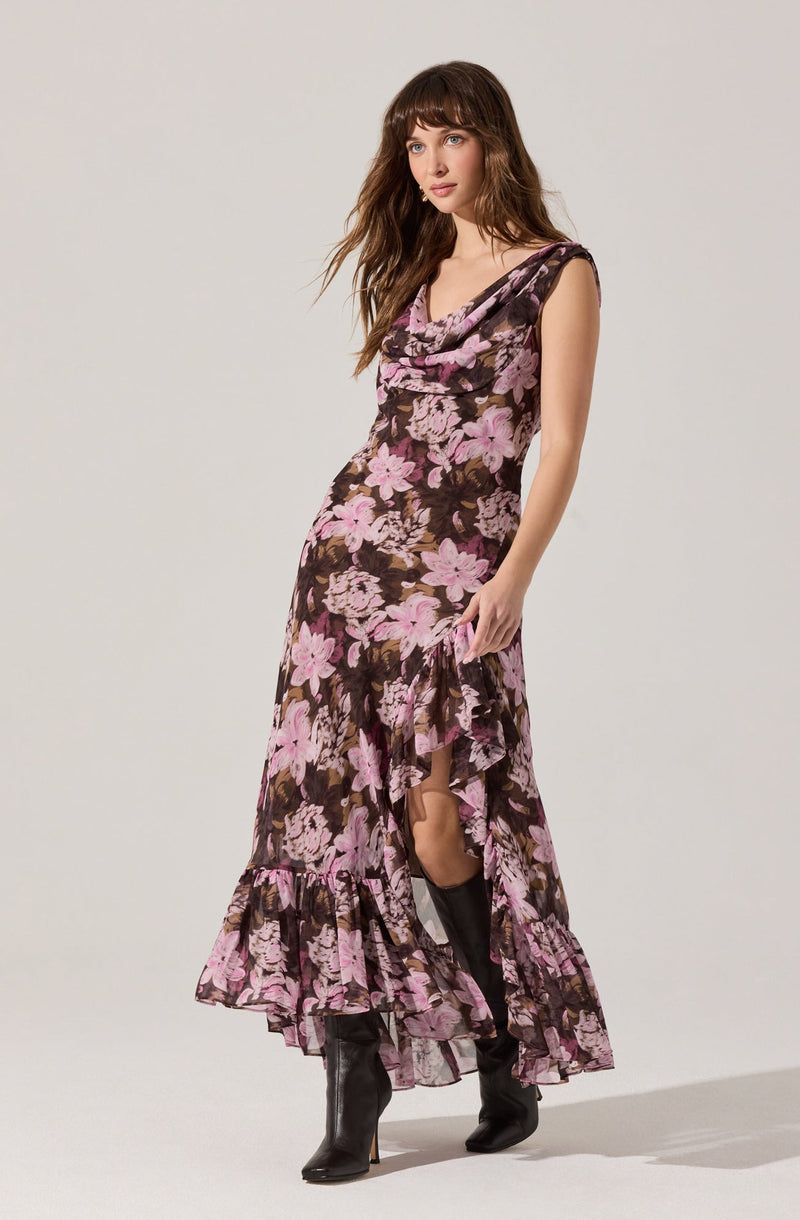 A woman is wearing a sleeveless, floor-length dress with a floral pattern in shades of pink and purple on a dark background. The dress features a cowl neckline, a fitted waist, and a ruffled hem with a slit that reveals her knee-high black boots.
