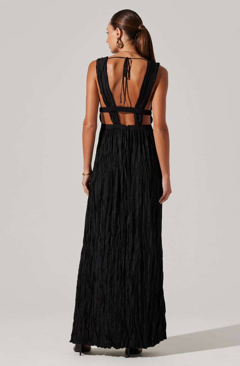 A woman is wearing a long, black dress with a textured, crinkled fabric. The dress features wide shoulder straps that extend over her shoulders and connect at the back with a tie closure. The back of the dress has cut-out sections, with two horizontal straps around the waist and a vertical open back with tied straps at the top. She is accessorized with large hoop earrings and a bracelet on her left wrist.
