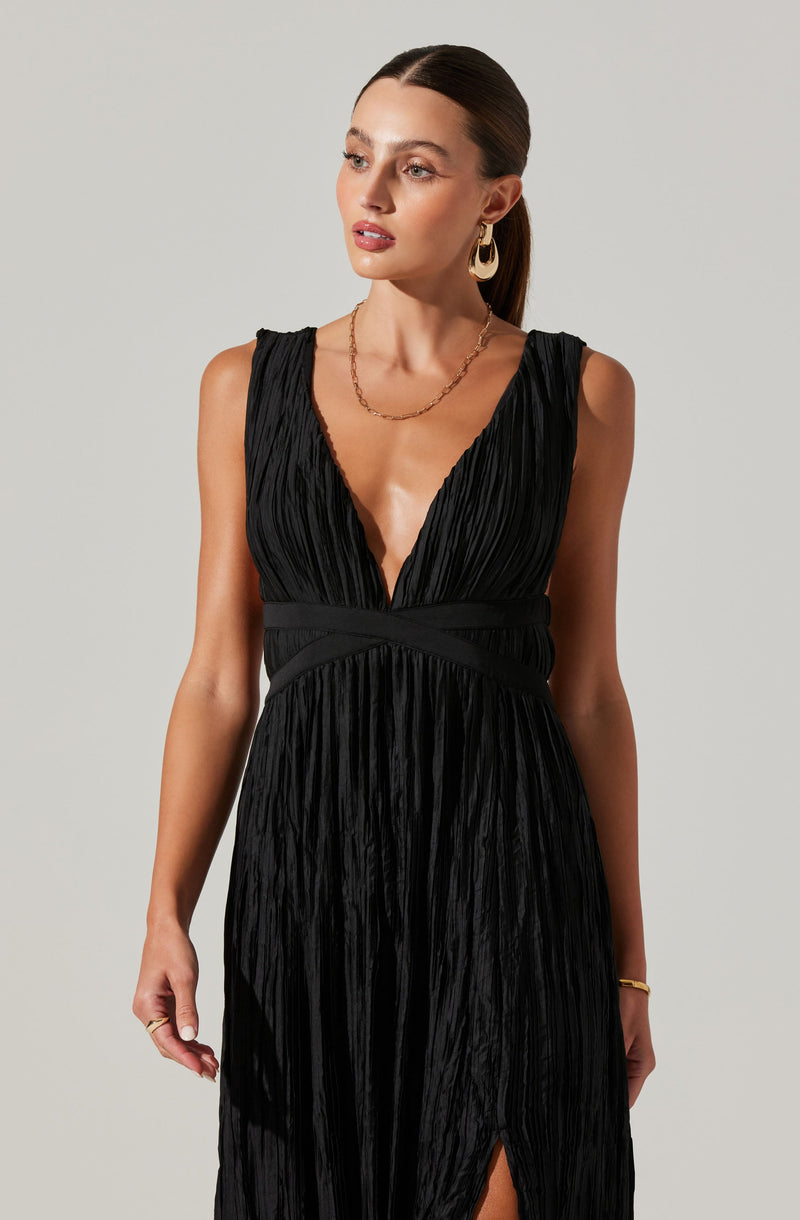 A woman is wearing a sleeveless black dress with a deep V neckline. The fabric has a shiny, crinkled texture, and the dress features a fitted waist with banded detailing. The skirt of the dress has a slit on one side. She accessorizes with large gold hoop earrings, a gold chain necklace, and a gold bracelet.