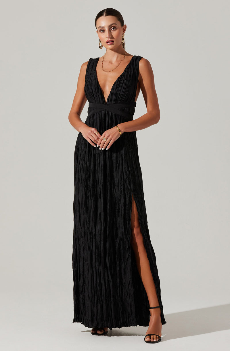 A woman is wearing a long, sleeveless black dress with a deep V neckline. The dress has a crinkled, textured fabric and features a high slit on one side, revealing her leg and a black ankle-strap high heel. She is accessorized with gold jewelry, including earrings, a necklace, and a bracelet.