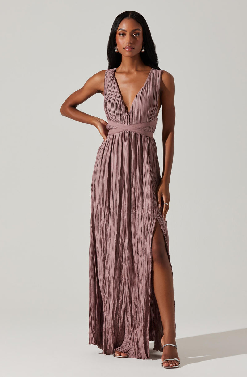 A woman is wearing a long, sleeveless dress in a shimmery mauve color. The dress features a deep V neckline with a textured, pleated fabric and has a fitted waistband that accentuates her waist. It also has a high slit on one side, revealing her leg and strappy high-heeled sandals.