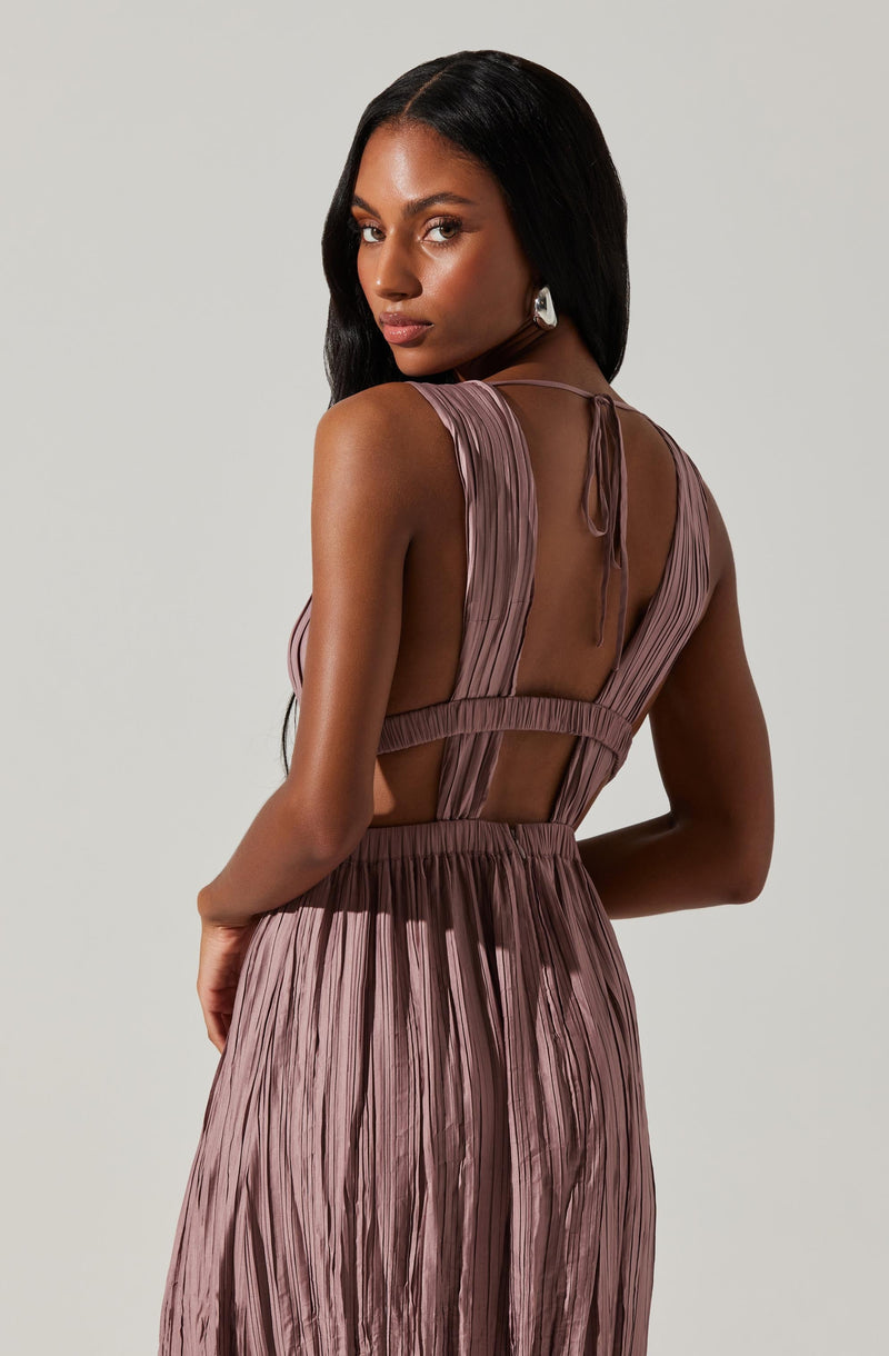 A woman is wearing a sleeveless, pleated, mauve-colored dress with sheer panels at the back and a tie closure at the neckline. The dress has a fitted waist and a flowing skirt with vertical pleats.