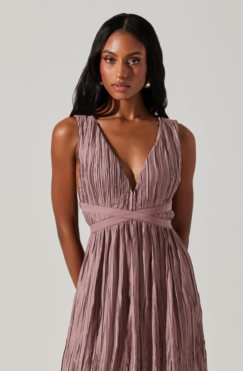A woman is wearing a sleeveless, deep V-neck dress with a pleated texture. The dress is a soft mauve or dusty rose color with a fitted waistband and a flowing skirt. The fabric has vertical pleats throughout, creating a textured appearance.