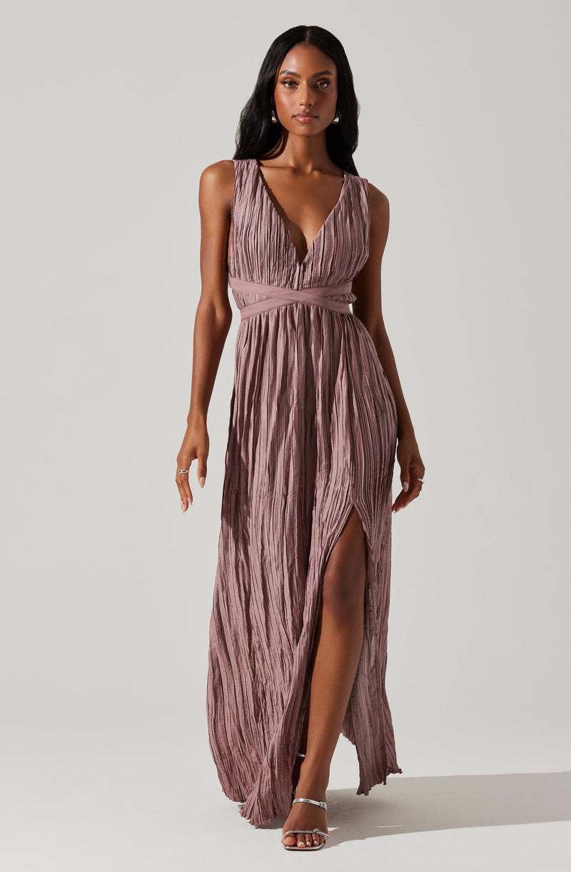 A woman is wearing a sleeveless, mauve-colored gown with a deep V-neckline and a pleated, textured fabric throughout. The dress features a fitted waistband and a high slit on the front, revealing her legs. She is also wearing high-heeled sandals.