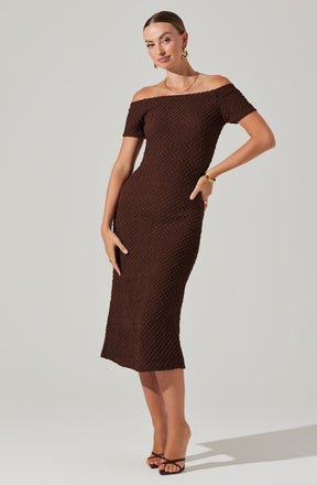 A woman is wearing a fitted, off-the-shoulder, short-sleeved, midi-length dress in a rich, dark brown color. The dress has a textured, woven pattern throughout. She is accessorized with large hoop earrings, a delicate necklace, a bracelet, and rings. She is also wearing strappy heeled sandals.