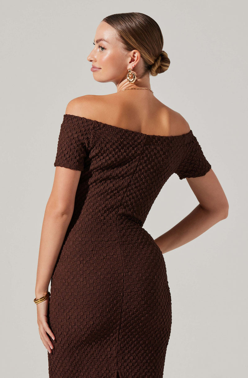 A woman is wearing a brown, textured, off-the-shoulder dress with short sleeves. The dress has a fitted silhouette, and she accessorizes with large, gold hoop earrings, a delicate chain necklace, and a bracelet. Her hair is styled in a sleek bun.