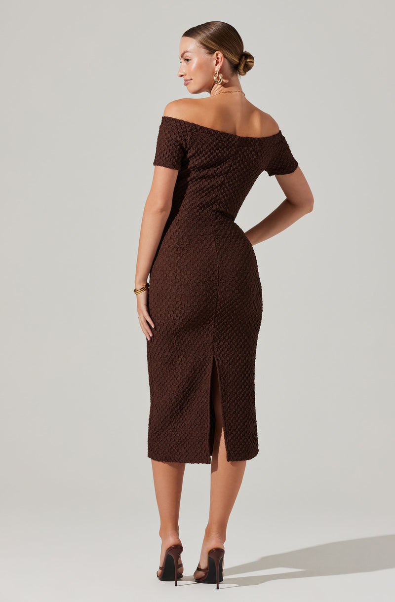 A woman is wearing a brown off-the-shoulder dress with a textured, honeycomb-like pattern. The dress is fitted and knee-length with a back slit. She is also wearing high-heeled shoes, gold earrings, and a gold bracelet. Her hair is styled in a low bun.