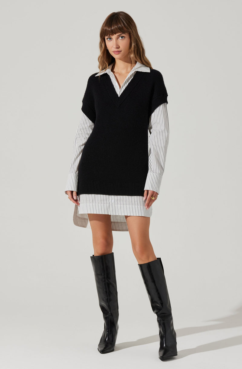 A woman is wearing a white shirt with thin black vertical stripes beneath a black sleeveless knit sweater vest. The shirt extends below the vest, creating a layered look, and the hem of the shirt is slightly longer in the back. She is also wearing a short, matching striped skirt and knee-high black patent leather boots.