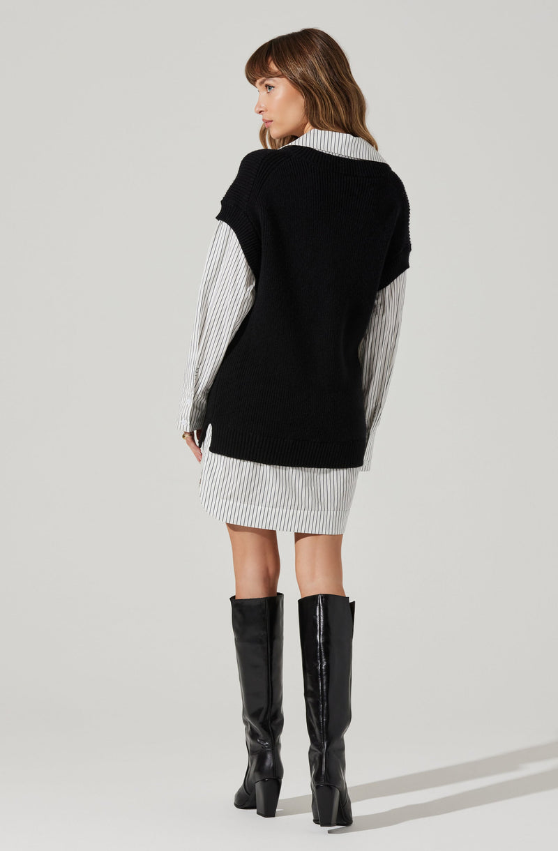 A woman is wearing a white dress with thin black vertical stripes, a black sleeveless knit sweater layered over the dress, and tall black patent leather boots with chunky heels.