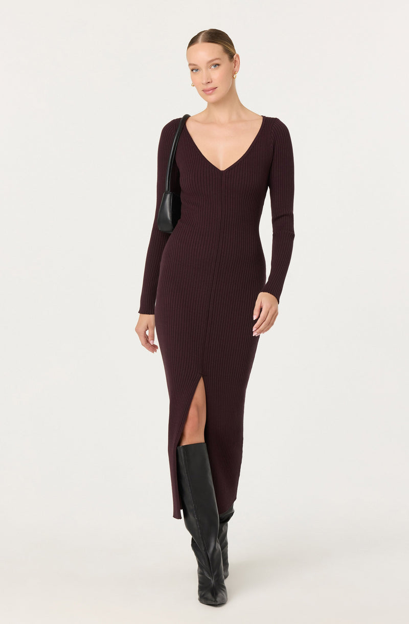 A woman is wearing a long, ribbed, burgundy dress with a deep V-neckline and long sleeves. The dress has a front slit that reaches mid-calf, revealing her black knee-high boots. She carries a black shoulder bag and has her hair styled in a sleek, pulled-back look.