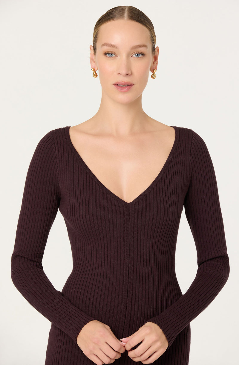 A woman is wearing a fitted, long-sleeved, deep burgundy ribbed knit dress with a V-neckline. She has gold hoop earrings and her hair is styled in a sleek bun.