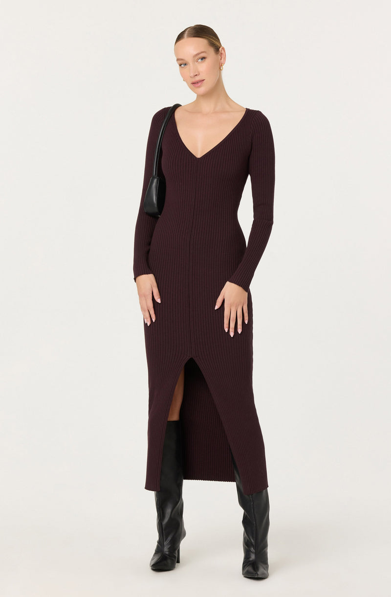A woman is wearing a long, fitted, ribbed knit dress in a deep burgundy color. The dress features a V-neckline, long sleeves, and a front slit that reveals a portion of her legs. She is also wearing knee-high black leather boots and carrying a black shoulder bag.