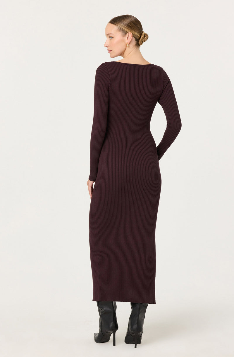 A woman is wearing a long, fitted, ribbed knit dress in a deep burgundy color. The dress has long sleeves and a round neckline. She is also wearing black high-heeled boots. Her hair is styled in a neat, low bun.