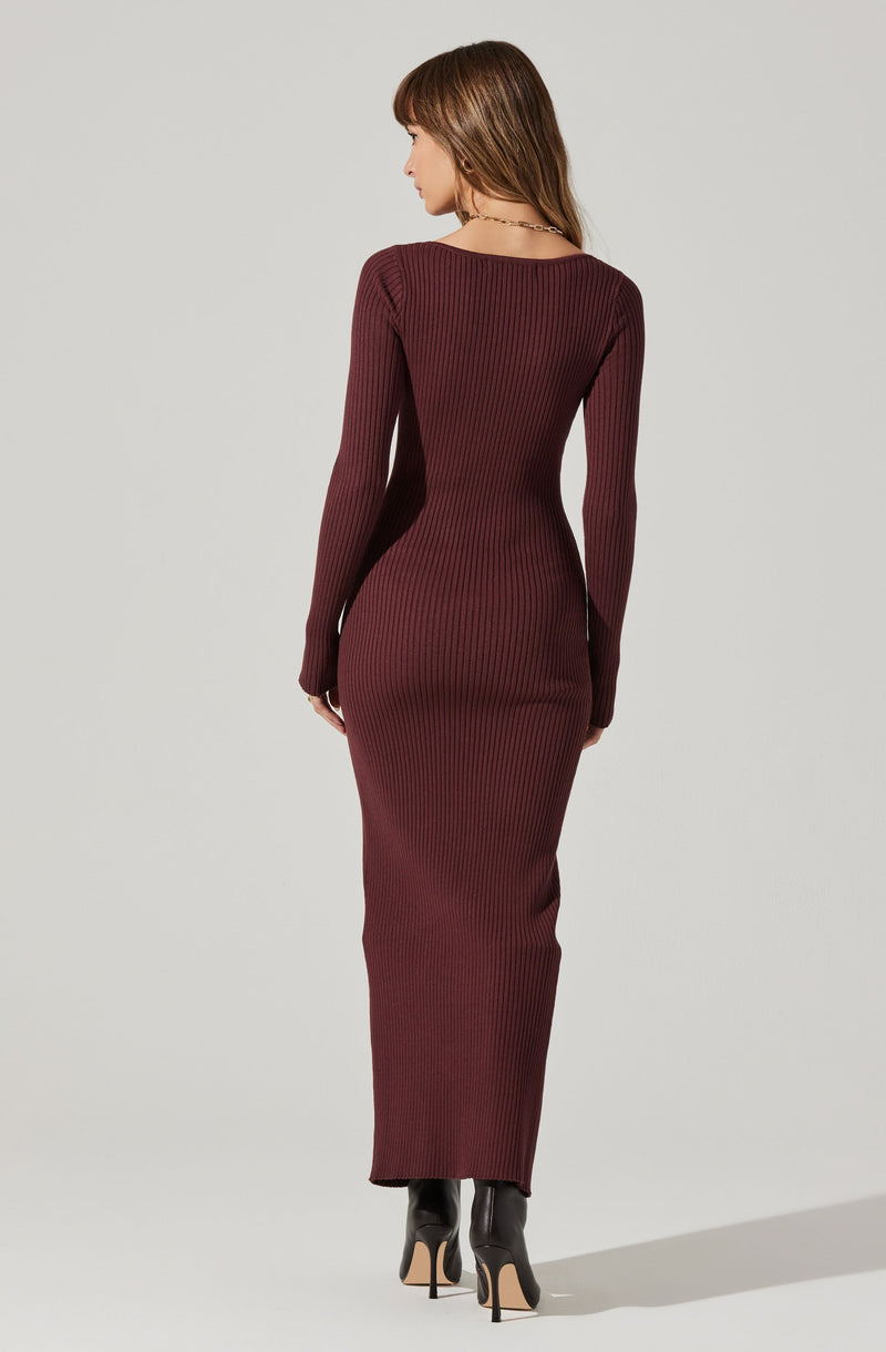 A woman is wearing a form-fitting, long-sleeved, ankle-length ribbed knit dress in a deep burgundy color. She has on black high-heeled ankle boots and is accessorized with a thin necklace. The dress features vertical ribbing and a round neckline. She is standing with her back facing the camera, and her hair is medium length and brown.