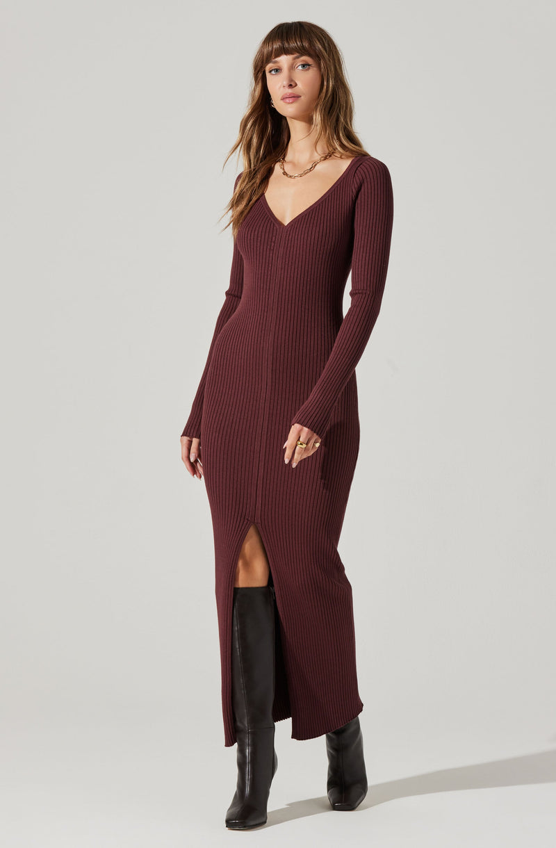 A woman is wearing a long, ribbed, burgundy knit dress with a V-neckline and long sleeves. The dress features a front slit that reveals her left leg and is paired with tall black leather boots. She is accessorized with a gold chain necklace and rings.