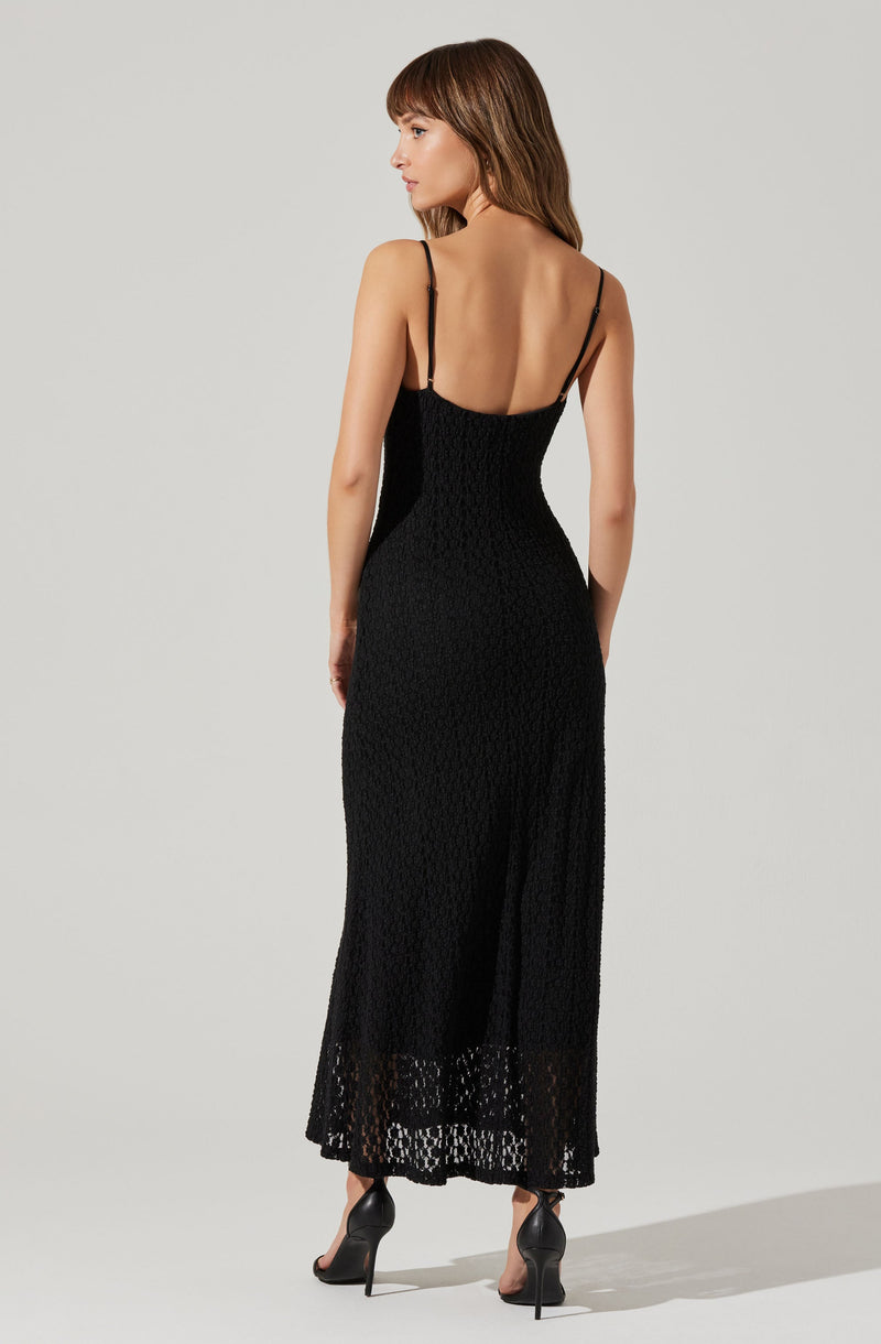 A woman is wearing a long black dress with thin spaghetti straps, fitted at the waist and hips, and featuring a textured, lace-like pattern throughout. The dress reaches down to her ankles with a slight flare at the hem. She is also wearing black high-heeled shoes.