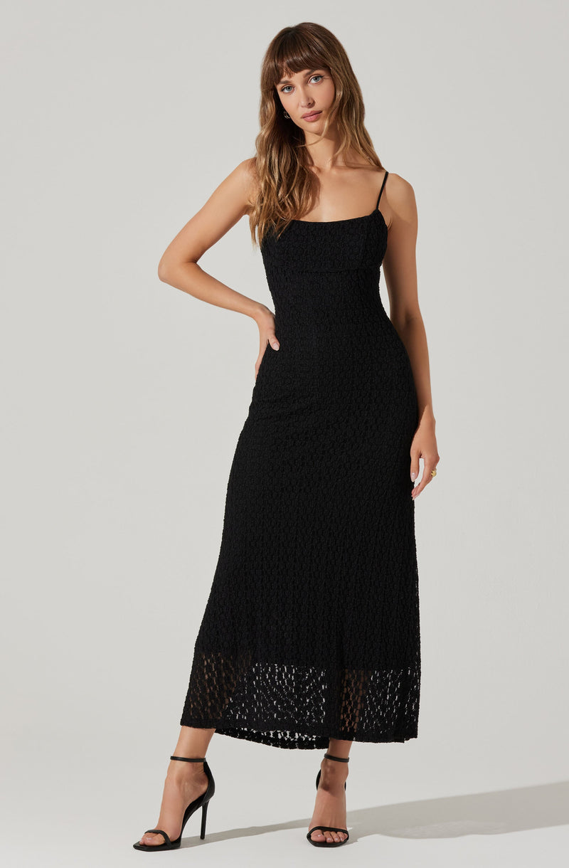 A woman is wearing a black, sleeveless, spaghetti strap dress with a fitted bodice. The dress features a textured, lace-like pattern with a semi-transparent hemline that extends to her ankles, revealing a lace detail at the bottom. She is also wearing black high-heeled sandals with thin straps around her ankles.