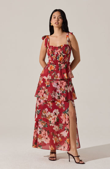 A woman is wearing a sleeveless, ankle-length dress with a floral pattern in shades of pink, red, orange, and green. The dress features shoulder ties, a square neckline with ruched bodice, and tiered ruffled layers with a slit on the side revealing her leg. She is also wearing black high-heeled sandals.
