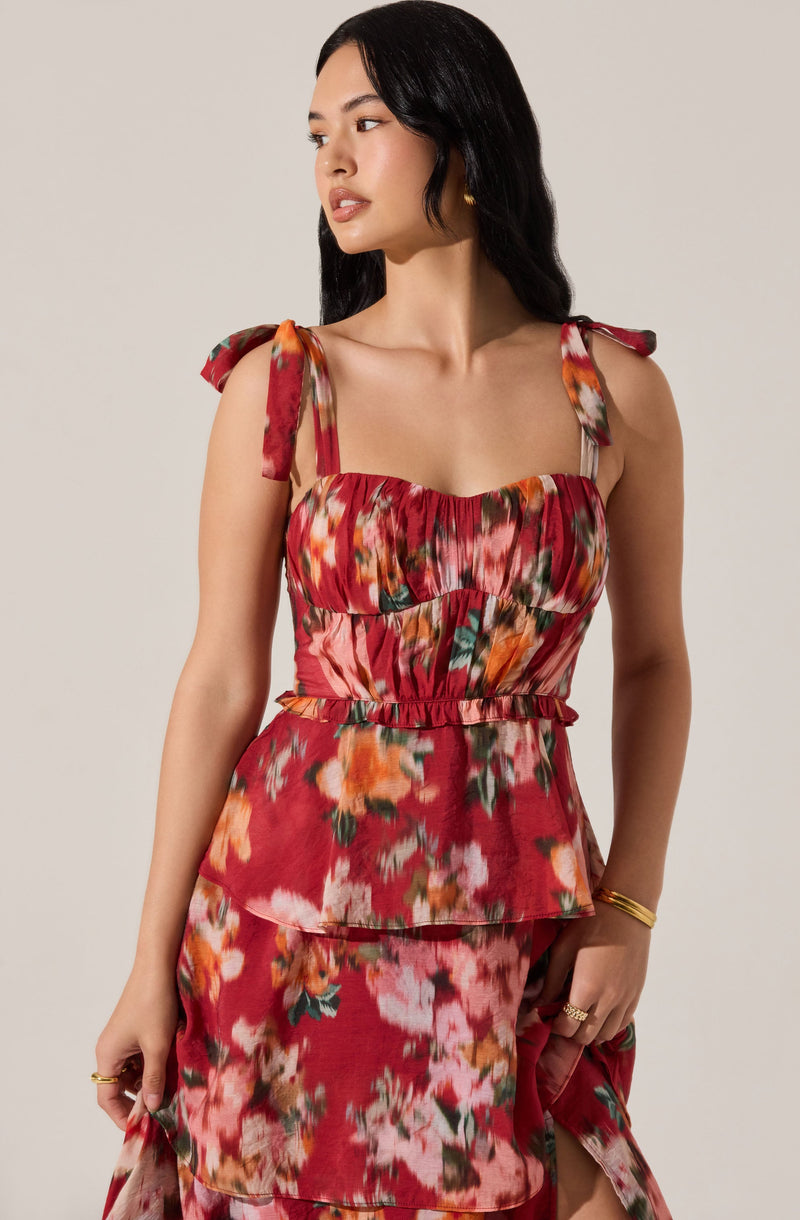 A woman is wearing a sleeveless, floral dress with a red background. The dress features decorative shoulder ties, a ruched bodice, and a tiered skirt with a slit. The fabric has a vibrant, multicolored floral pattern with shades of pink, orange, green, and white.