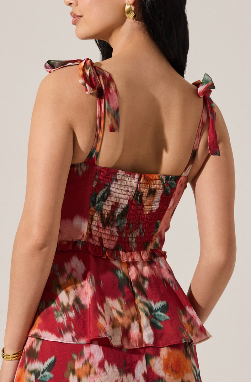 A woman is wearing a sleeveless dress with shoulder ties. The dress has a floral pattern in shades of red, pink, orange, and green. The top part of the dress is fitted with shirring detail, and the skirt has a layered, ruffled design.