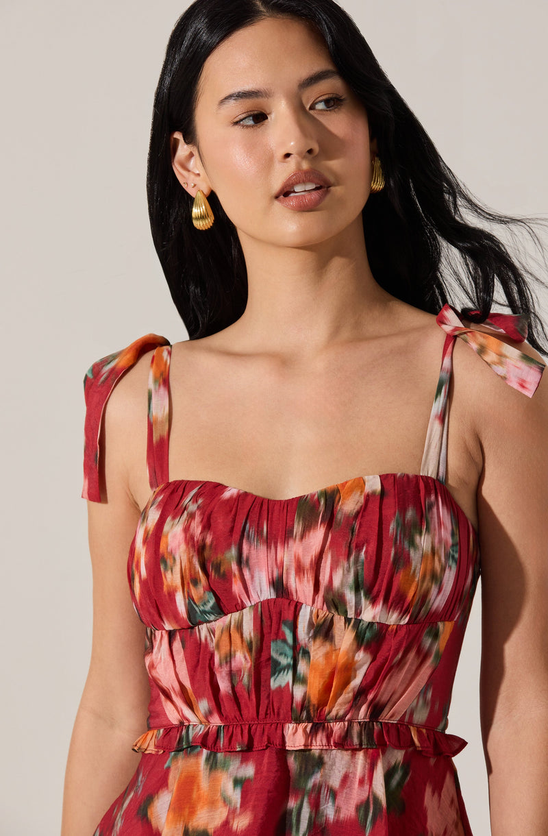 A woman is wearing a sleeveless dress with shoulder straps tied into bows on each shoulder. The dress features a colorful, abstract print in shades of red, orange, pink, green, and white, with a gathered, ruched bodice and a ruffled waistband. She has long, dark hair and is accessorized with large, gold, shell-shaped earrings.