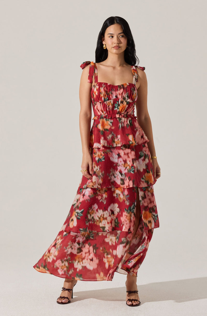 A woman is wearing a sleeveless, tiered maxi dress with a floral pattern in shades of red, pink, orange, and green. The dress has shoulder ties and a smocked bodice. She is accessorizing with gold hoop earrings, a gold bracelet, and black strappy heels.