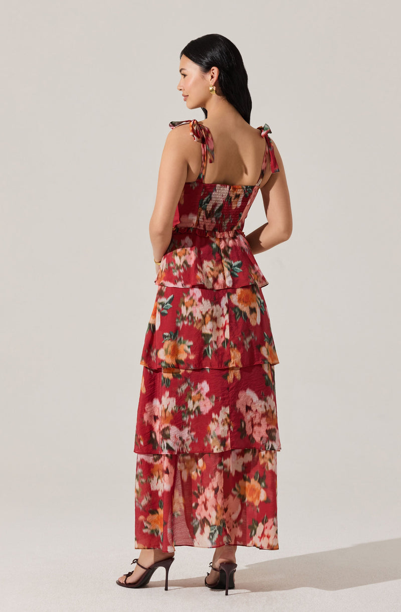 A woman is wearing a floral printed dress with multiple tiers and ruffle details. The dress has a fitted bodice with shoulder straps tied into bows. The fabric features a colorful floral pattern with shades of pink, red, orange, green, and white. She is accessorizing with gold earrings and wearing black high-heeled sandals.