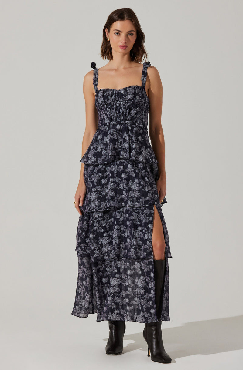 The dress is a sleeveless, ankle-length garment with a dark base color and a light floral print. It features adjustable shoulder straps tied into bows, a fitted bodice, and a tiered, slightly ruffled skirt with a high slit on one side. The fabric appears lightweight and semi-sheer.