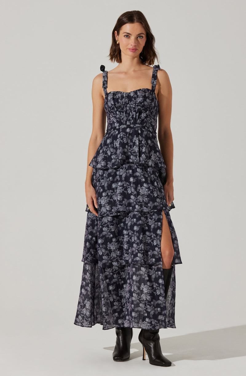 A woman is wearing a sleeveless, dark-colored dress with a floral pattern. The dress features tie straps at the shoulders, a fitted smocked bodice, and a tiered skirt with a thigh-high slit on one side. She is also wearing black high-heeled boots.