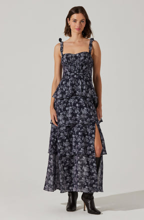 A woman is wearing a sleeveless, dark-colored dress with a floral pattern. The dress features tie straps at the shoulders, a fitted smocked bodice, and a tiered skirt with a thigh-high slit on one side. She is also wearing black high-heeled boots.