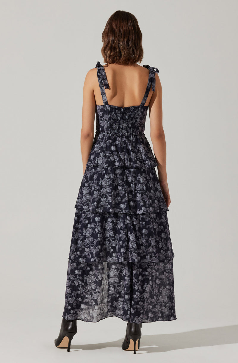The dress is a sleeveless, full-length gown with a black background and a small, light-colored floral pattern. It features shoulder straps tied into bows at the shoulders and a fitted bodice with a gathered waist. The skirt has multiple tiers or ruffles, adding volume and movement to the dress. The fabric appears lightweight and slightly sheer.