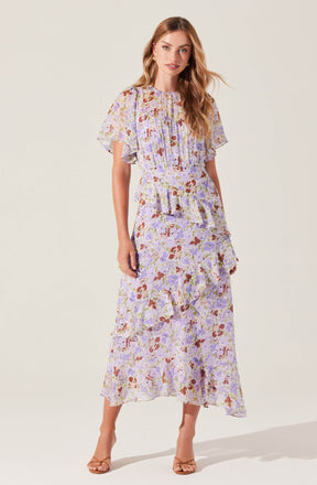 A woman is wearing a long, flowy dress with short, loose sleeves. The dress features a floral pattern in shades of purple, lavender, brown, and cream. It has a high, round neckline and is fitted at the waist with a ruffled detail, cascading into a tiered, ruffled skirt that reaches down to her ankles. She is also wearing nude-colored high-heeled sandals.