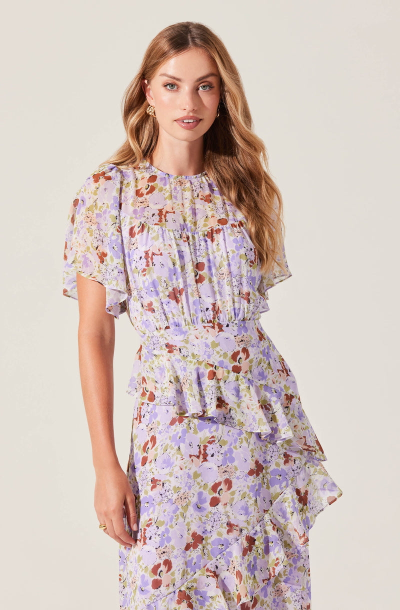 A woman is wearing a light-colored, floral print dress with shades of purple, lavender, brown, and cream. The dress features a round neckline, short flutter sleeves, and a fitted waist. The skirt has multiple ruffled layers that cascade down, creating a flowing silhouette.