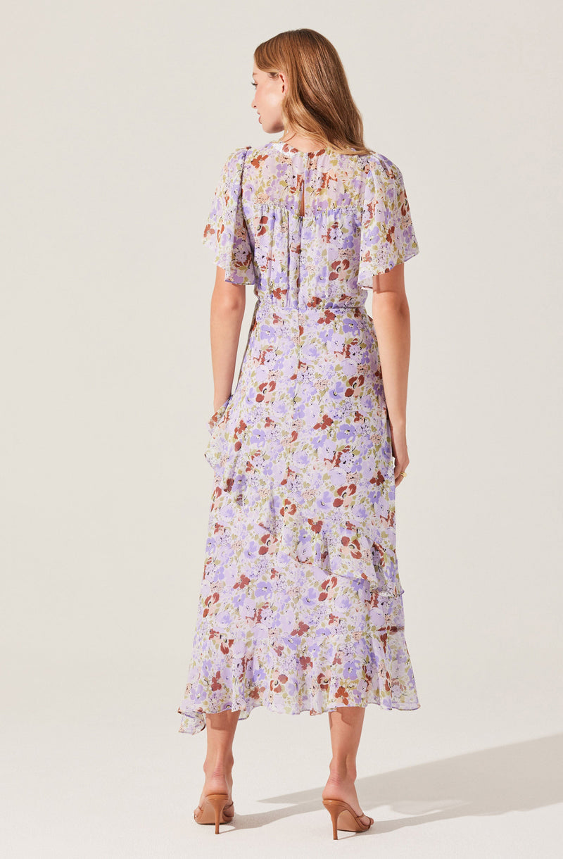 A woman is wearing a light-colored, floral-print dress with short, loose sleeves and a ruffled hem. The dress has a flowing, midi length and features a tiered design with ruffles along the skirt. The fabric displays a pattern of purple, pink, orange, and green flowers.