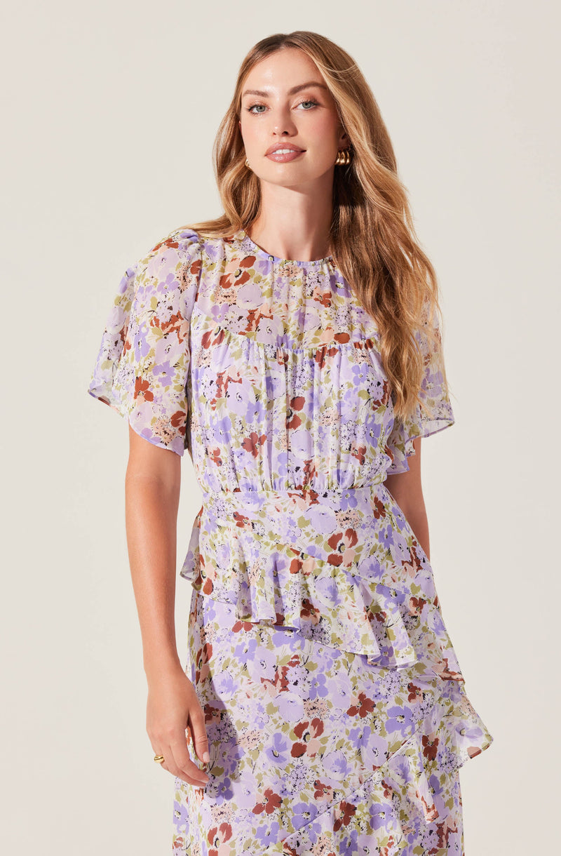 A woman is wearing a matching floral outfit made of lightweight, semi-sheer fabric. The top features short, puffed sleeves and a round neckline, with a loose fit. The skirt has a tiered, ruffled design with a flowing silhouette, and the pattern consists of abstract floral prints in shades of lavender, purple, reddish-brown, and white.