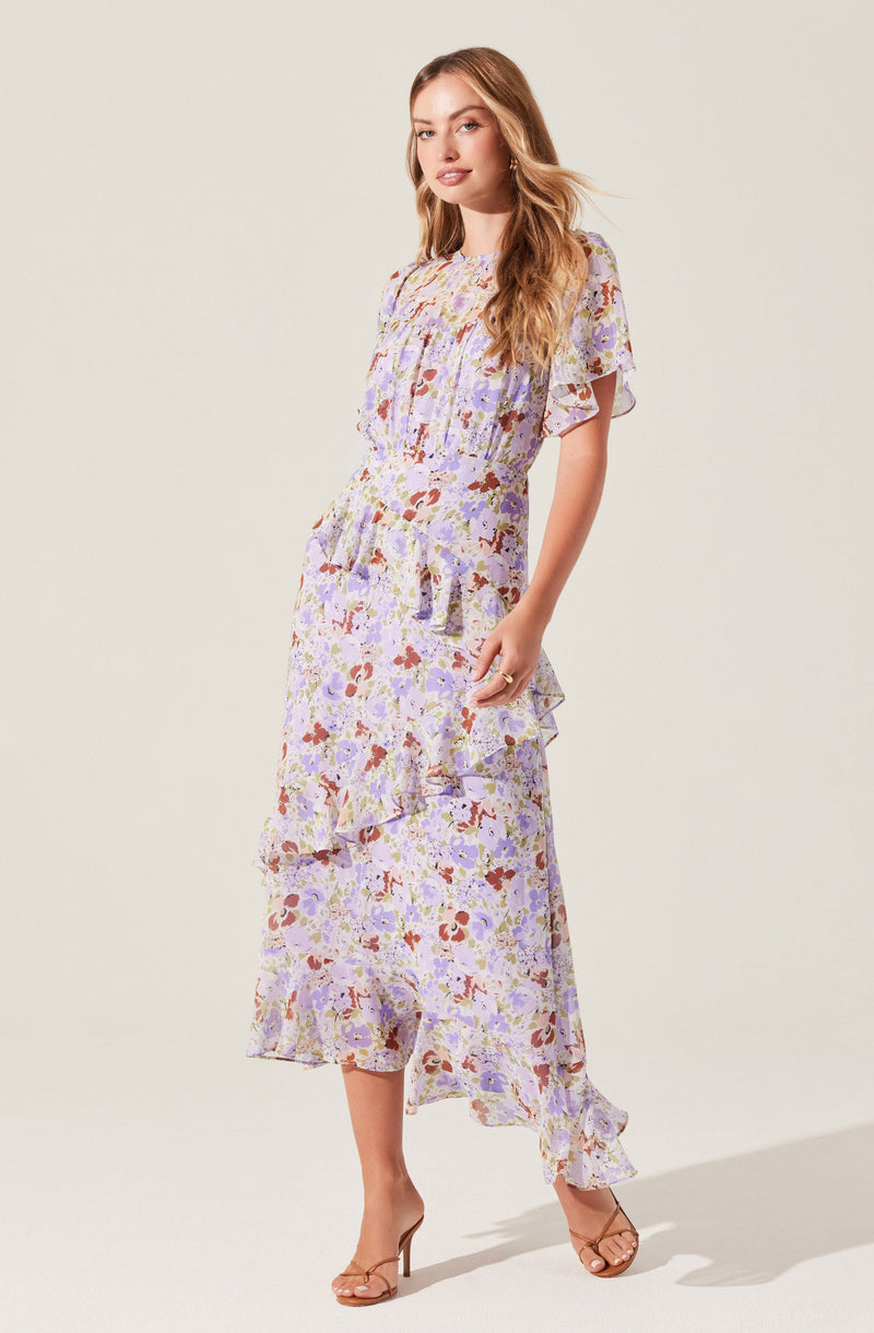 A woman is wearing a long, flowy maxi dress with a floral pattern in shades of purple, lavender, red, and white. The dress features short, flutter sleeves, a cinched waist, and a ruffled hemline that adds a layered detail to the skirt. She is wearing nude, strappy high-heeled sandals.