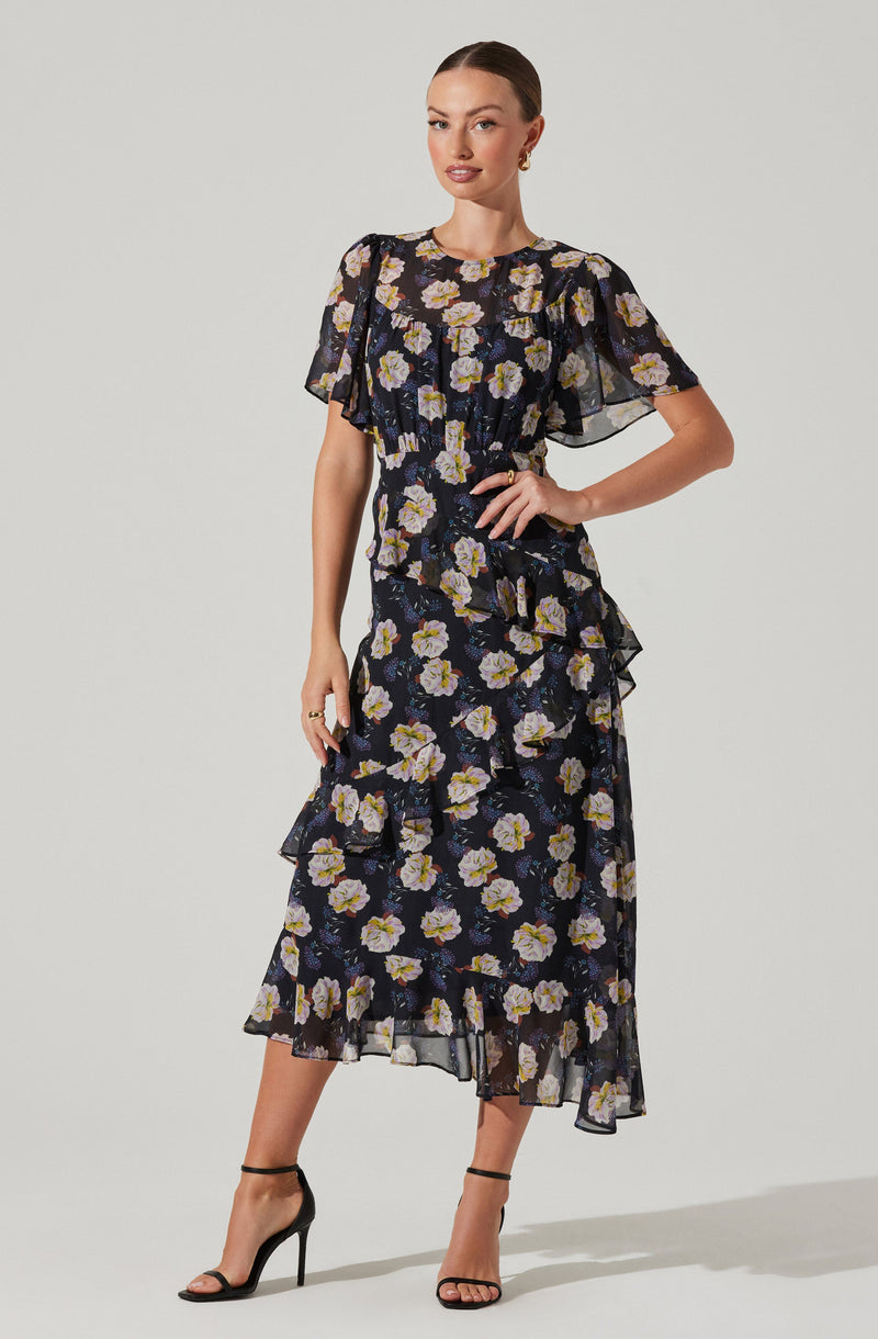 A woman is wearing a two-piece dress set with a floral pattern. The top is a short-sleeved, black sheer fabric with large, light-colored flowers and ruffle details on the sleeves and around the waist. The skirt is a matching black sheer fabric with the same large floral pattern, featuring ruffles along the layers and an asymmetrical hemline. She is also wearing black high-heeled sandals with ankle straps.