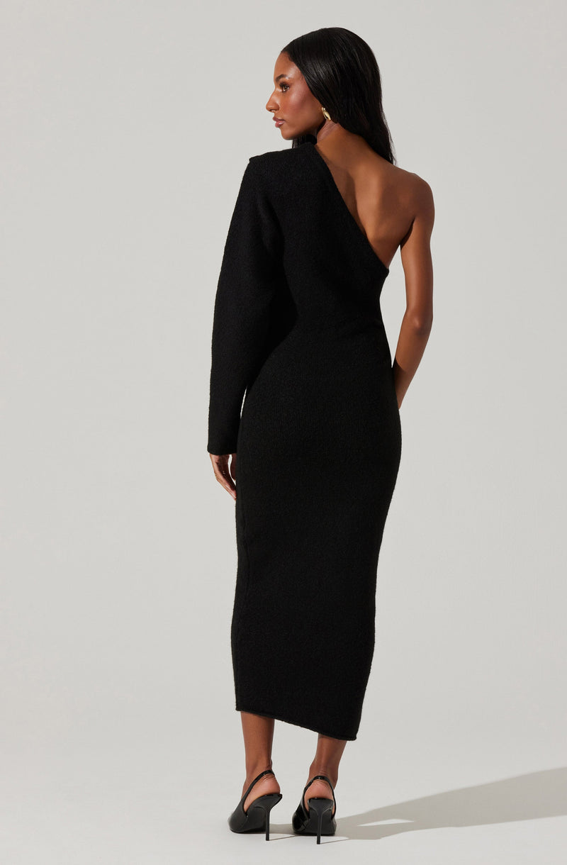 A woman is wearing a form-fitting, black dress with an asymmetrical design. The dress features a single long sleeve on the left arm, while the right side is sleeveless, exposing her shoulder and upper back. She is also wearing black high-heeled shoes.