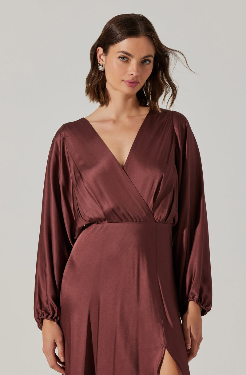 A woman is wearing a satin dress in a deep burgundy color. The dress features a V-neckline with a wrap style at the top, long puffed sleeves gathered at the cuffs, and a high slit on the skirt.
