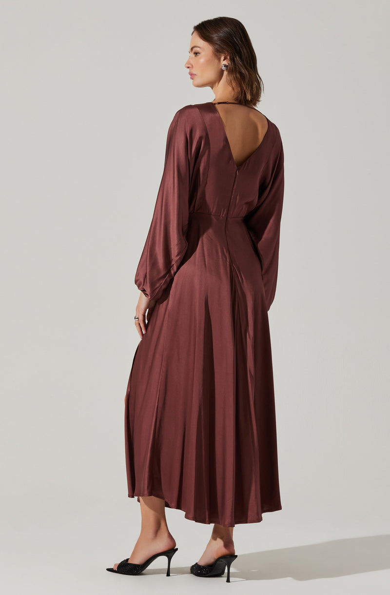 A woman is wearing a long, satin, burgundy dress with long billowy sleeves that gather at the wrists. The dress has a deep V-shaped back neckline and a fitted waist, flowing into a pleated, ankle-length skirt. She is paired with black high-heeled sandals that have thin straps.