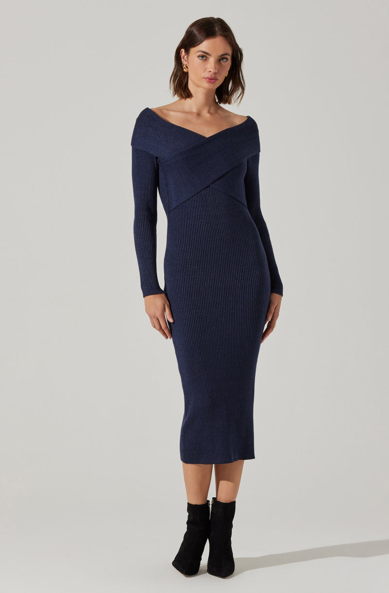 A woman is wearing a fitted, ribbed navy blue knit dress with an off-the-shoulder neckline that wraps around her shoulders and crosses at the front. The dress has long sleeves and reaches below the knees. She is also wearing black ankle boots.