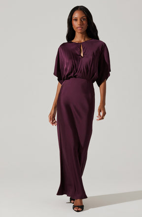 A woman is wearing a deep purple satin dress with a loose-fitting, draped top that has wide sleeves and a keyhole neckline, paired with a fitted, high-waisted, long skirt that reaches the floor. She is also wearing black open-toe heels.
