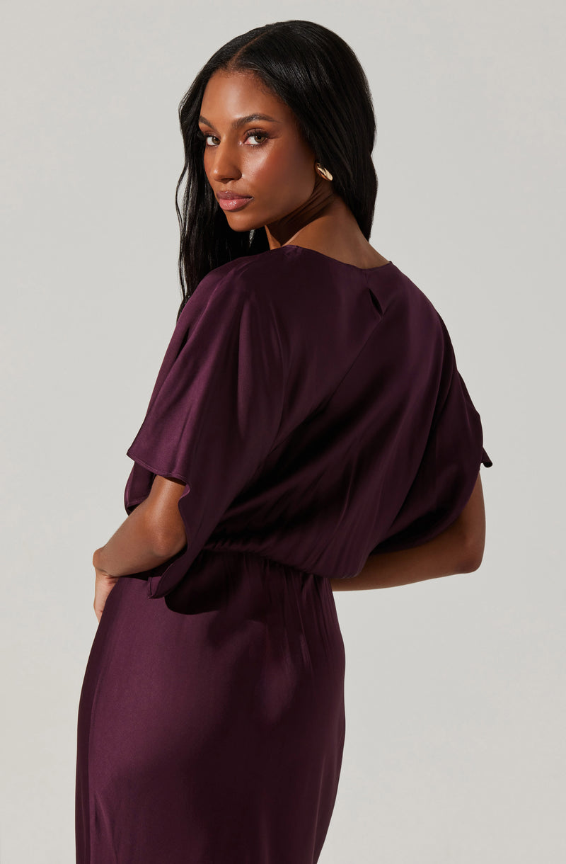 A woman is wearing a deep purple, silky dress with a loose-fitting top that has short, puffed sleeves. The dress has a gathered waistline, creating a smooth, flowing silhouette.