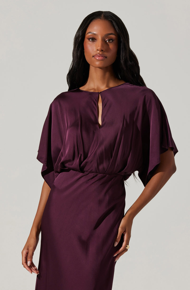 A woman is wearing a deep purple satin dress with a loose-fitting top that has draped, flowing sleeves and a keyhole opening at the neckline. The dress has a fitted waist and a smooth, flowing skirt made of matching satin fabric.